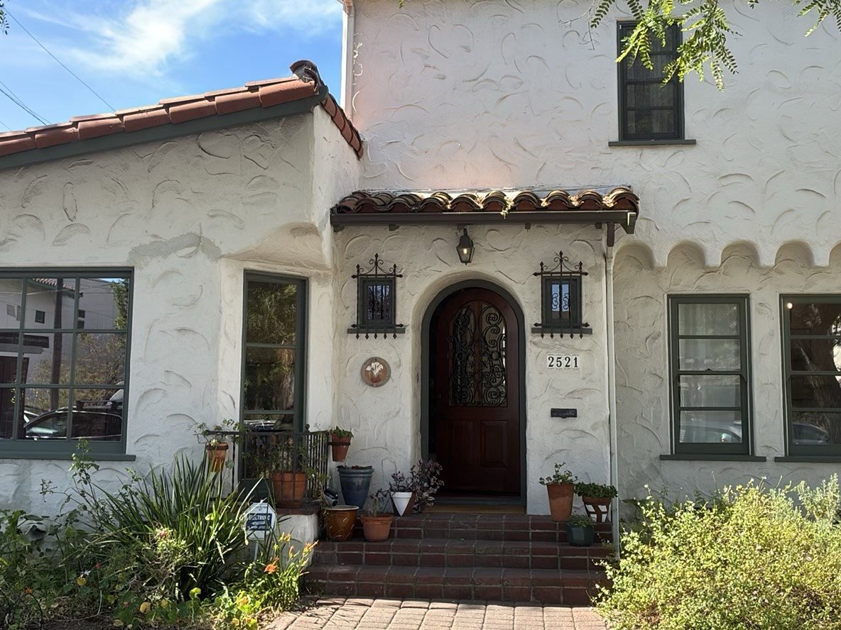 Front exterior of a Spanish-style stucco house with terracotta tile roof, arched entry door, and manicured landscaping in San Diego