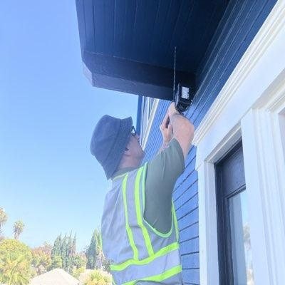 Inspector in safety vest pressing a FLIR thermal imaging device directly against an exterior soffit panel from a ladder position
