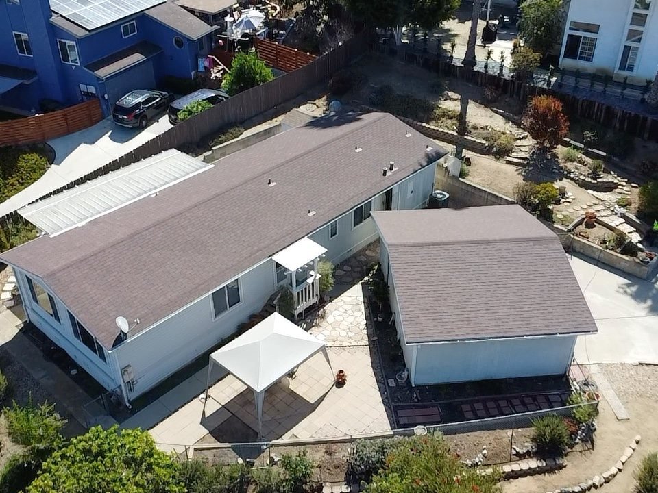 Drone photograph looking straight down at a large low-slope asphalt shingle roof covering a substantial building footprint, showing multiple roof sections, a covered patio, and surrounding neighborhood