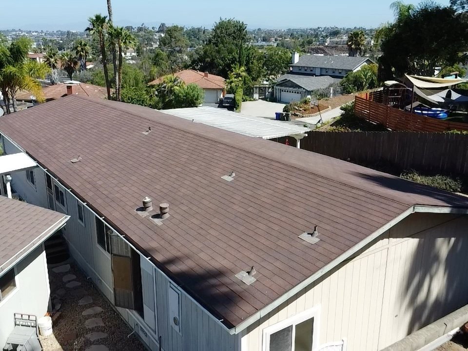 Angled drone view over a wide brown asphalt shingle roof showing vent pipes and roof vents, with palm trees, homes, and the San Diego skyline visible in the distance under a blue sky