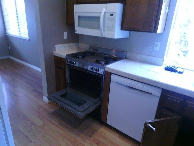 Kitchen overview showing warm wood upper and lower cabinets, hardwood floors, natural light from large windows, and open flow into adjacent living room