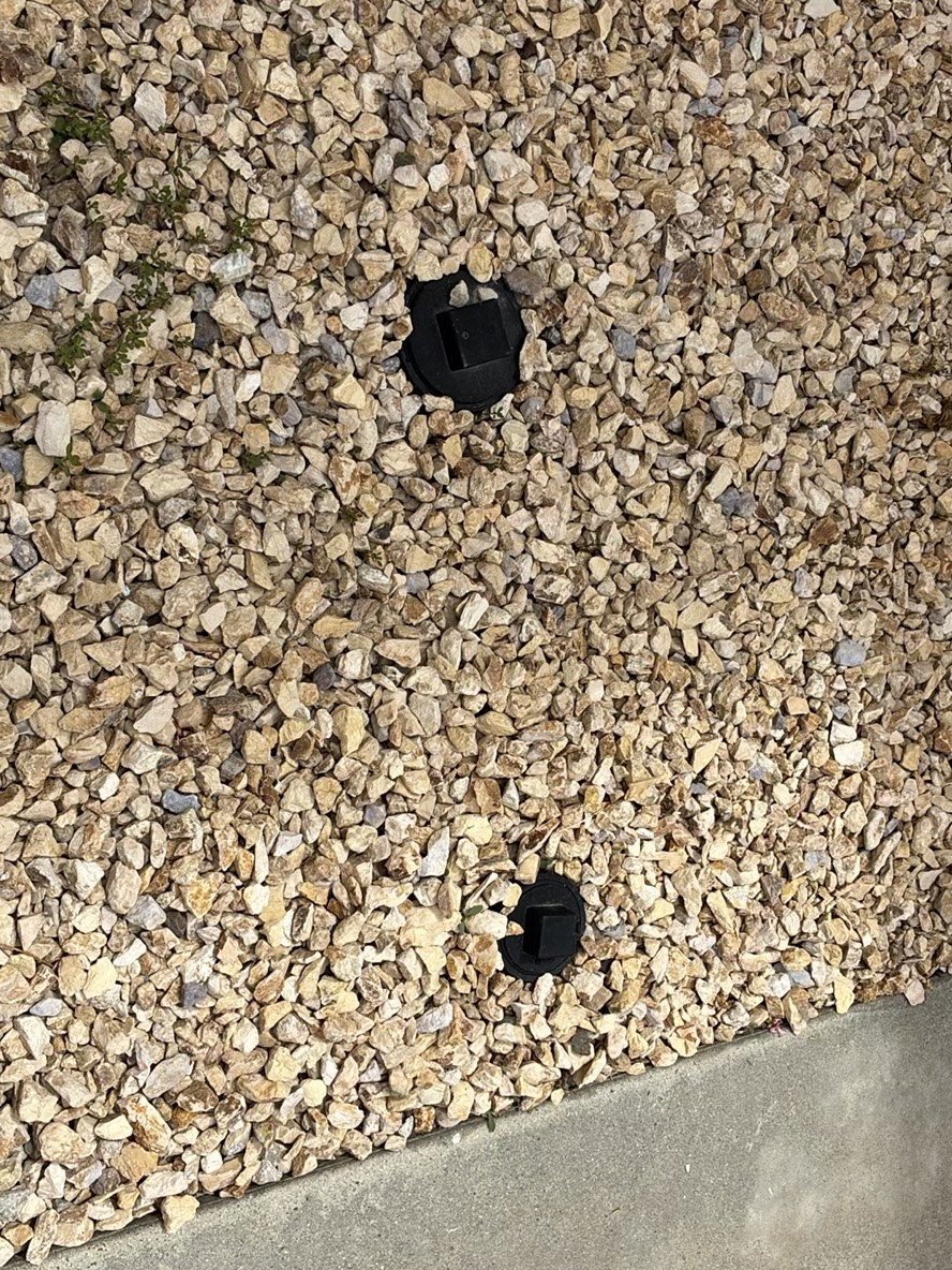 Two black sewer cleanout caps flush with gravel landscaping next to a concrete foundation, providing drain access points for sewer scope inspection