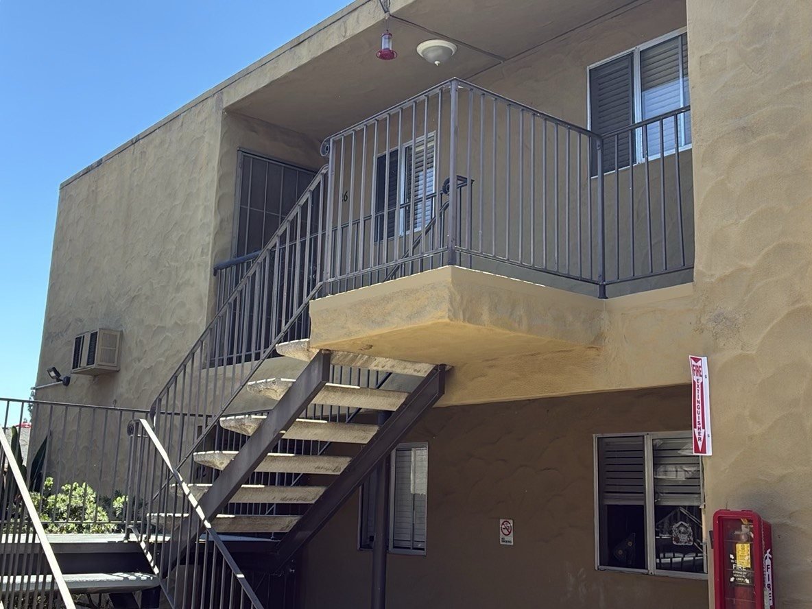 Two-story stucco apartment building with an exterior steel staircase leading to an upper-level stair landing enclosed by steel guardrails, fire extinguisher visible near ground-level units.