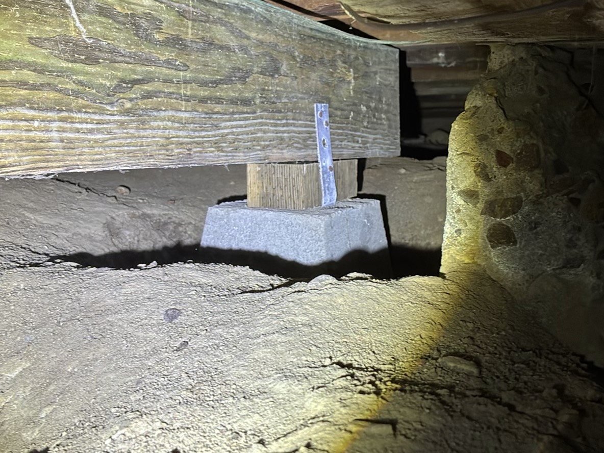 Close-up view inside a crawl space showing a concrete pad footing supporting a wood post and floor framing, documented during a foundation inspection