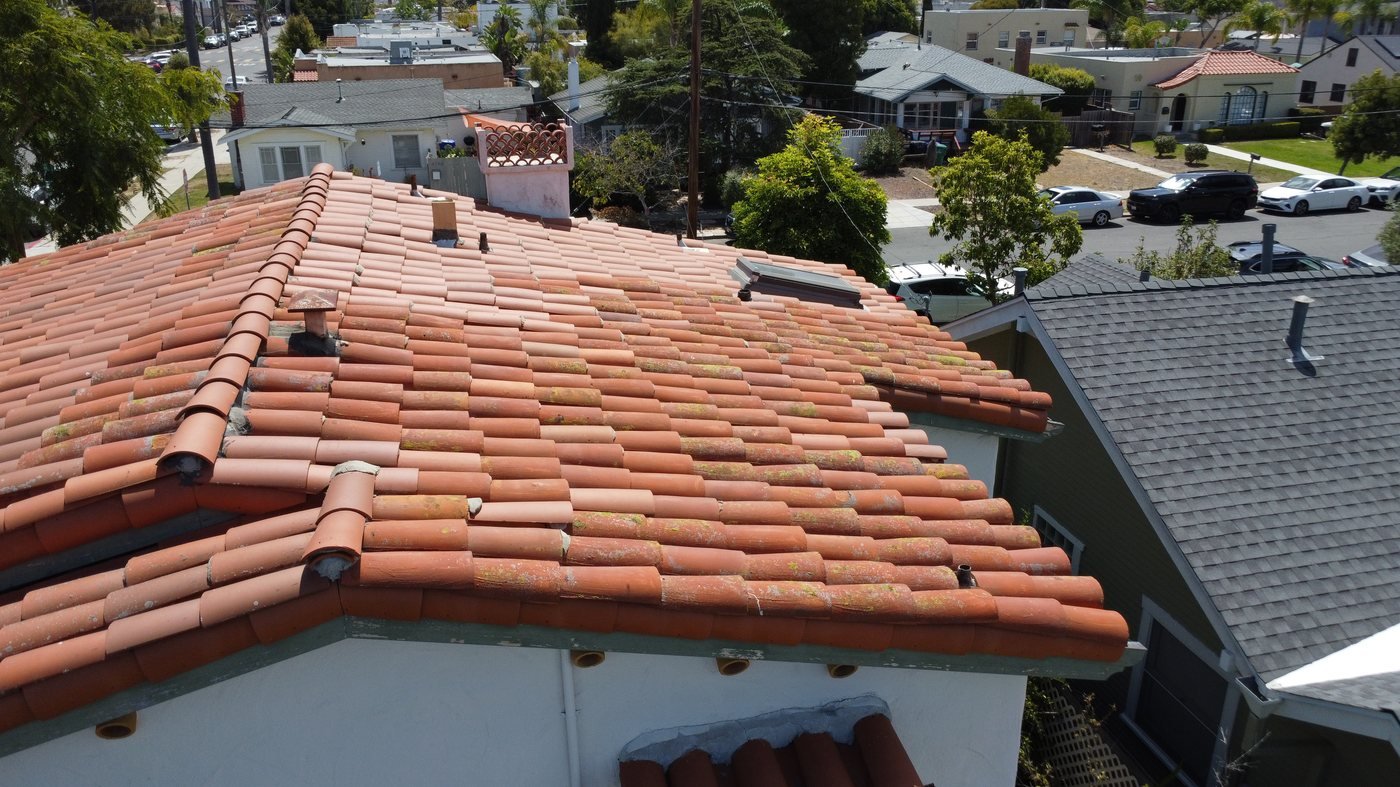 Elevated perspective looking across a clay barrel tile roof showing the chimney cap, plumbing vent pipes, and neighboring homes and street below