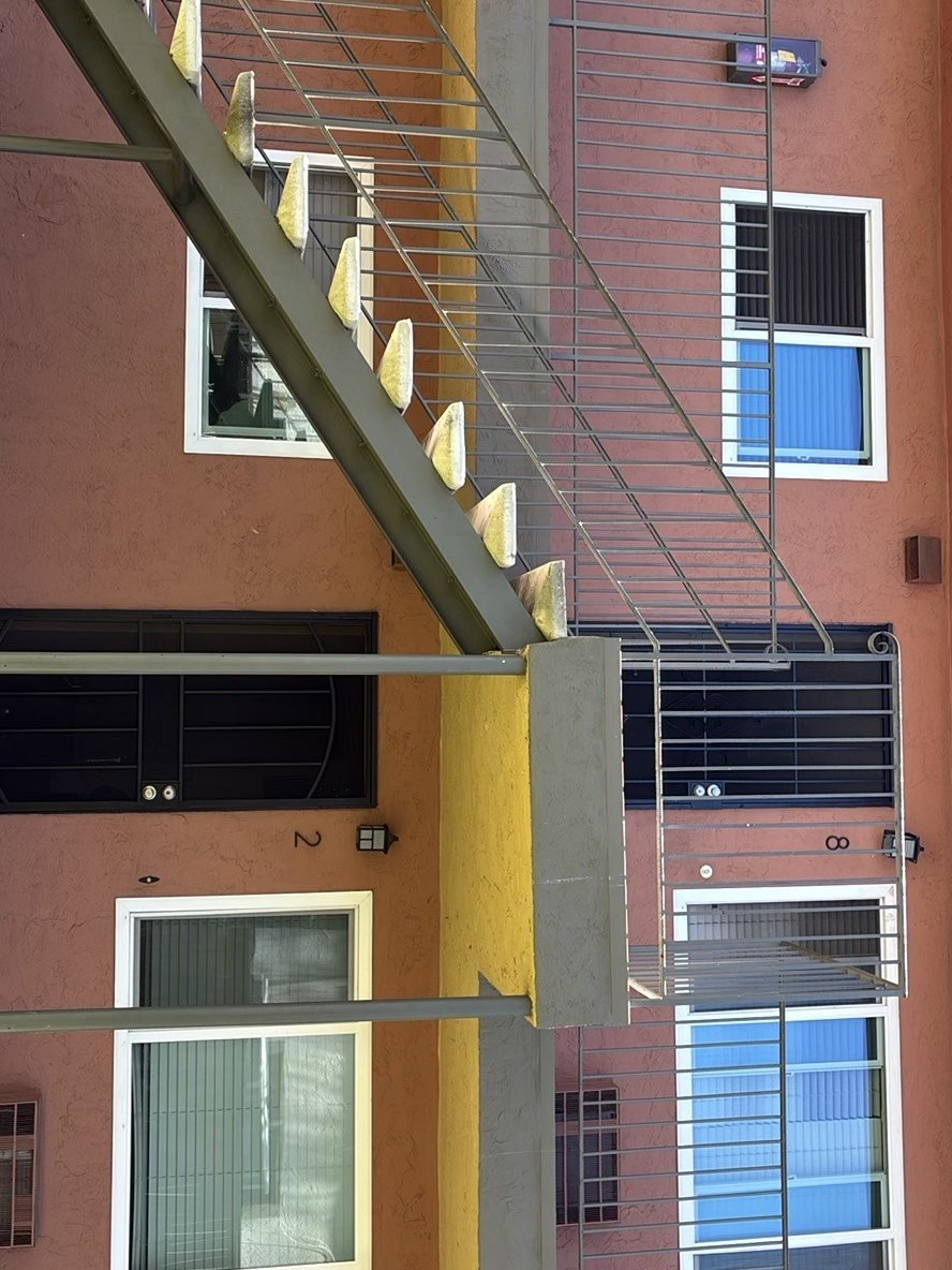 Close-up of an exterior steel staircase at a multi-unit apartment building showing angled stair stringers, metal guardrails, and unit numbers 2 and 8 on terracotta-stucco walls.