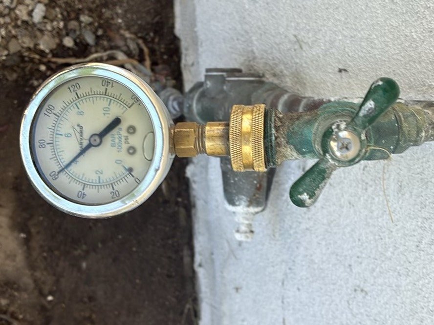Water pressure gauge with brass connector attached to a green outdoor hose bib mounted on a stucco wall, used to test residential water pressure during inspection