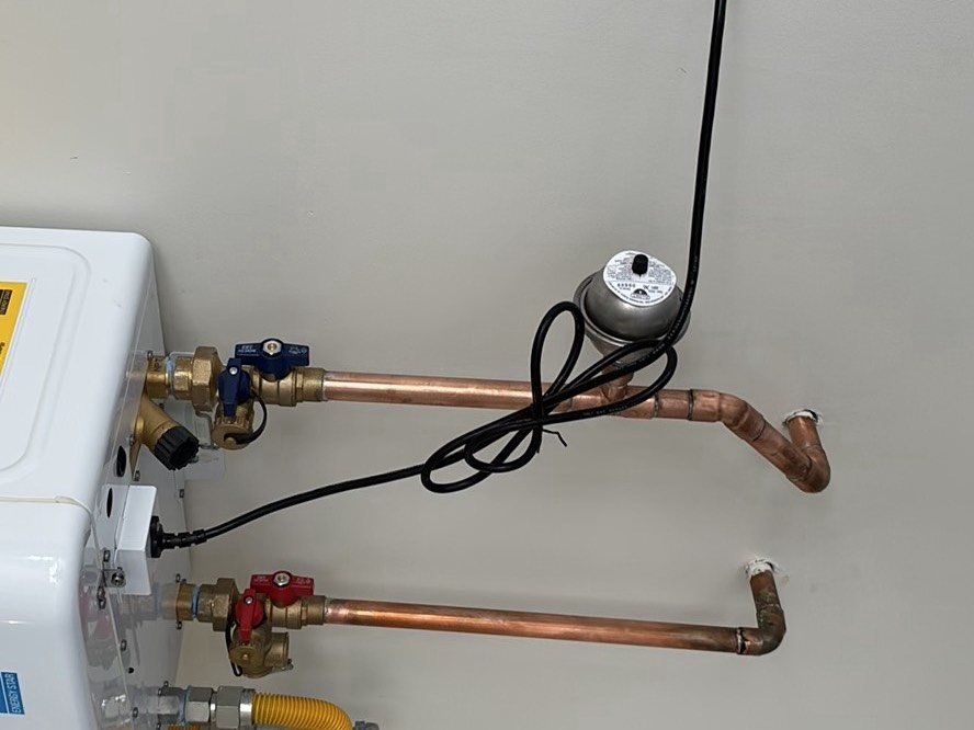 Close-up of copper water supply pipes with brass valves, a recirculating pump, and black wiring attached to a water heater during a home inspection