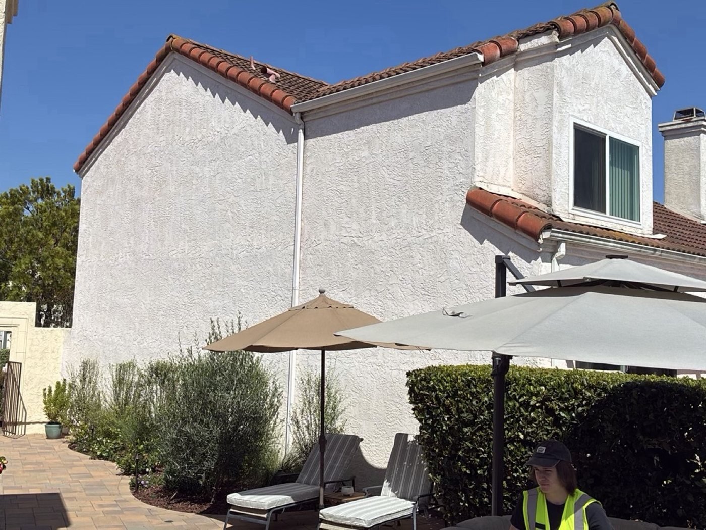 Inspector in yellow safety vest standing at the rear exterior of a two-story stucco home with concrete paver patio visible at the foundation base level