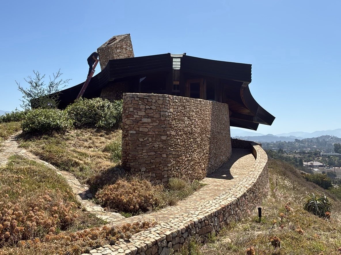 Inspector climbing a ladder against the exterior stone wall of a unique hillside property to access the flat roof above, surrounded by native vegetation and San Diego hills