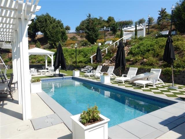 Rectangular swimming pool with white pergola, multiple lounge chairs, manicured landscaping, and a terraced hillside garden in the background