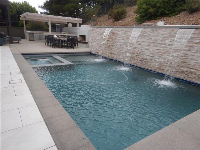 Contemporary rectangular swimming pool with raised spa, cascading stone waterfall wall, and a covered outdoor dining and kitchen area