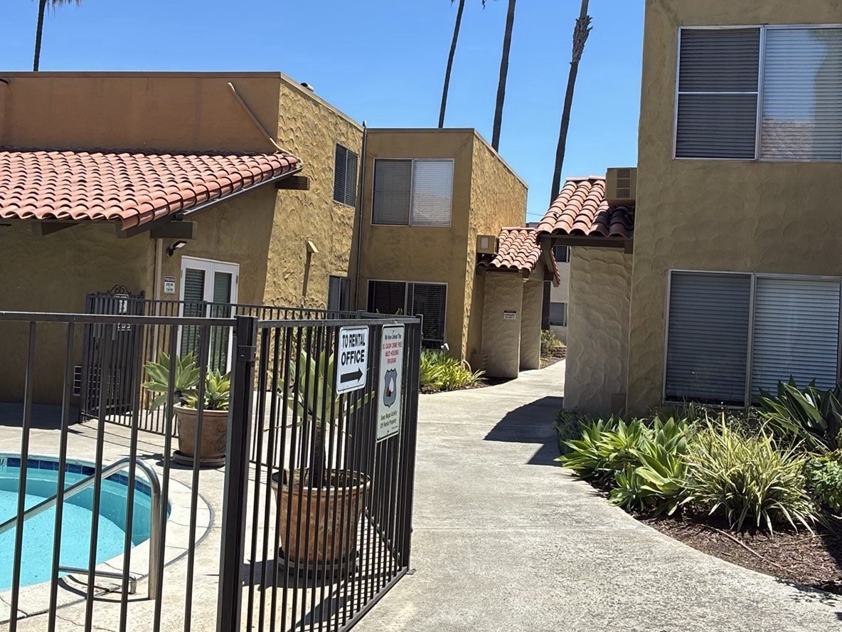 Attractive two-story Spanish-style apartment complex with terracotta tile roofs, palm trees, a gated pool area, and a well-maintained walkway on a sunny day