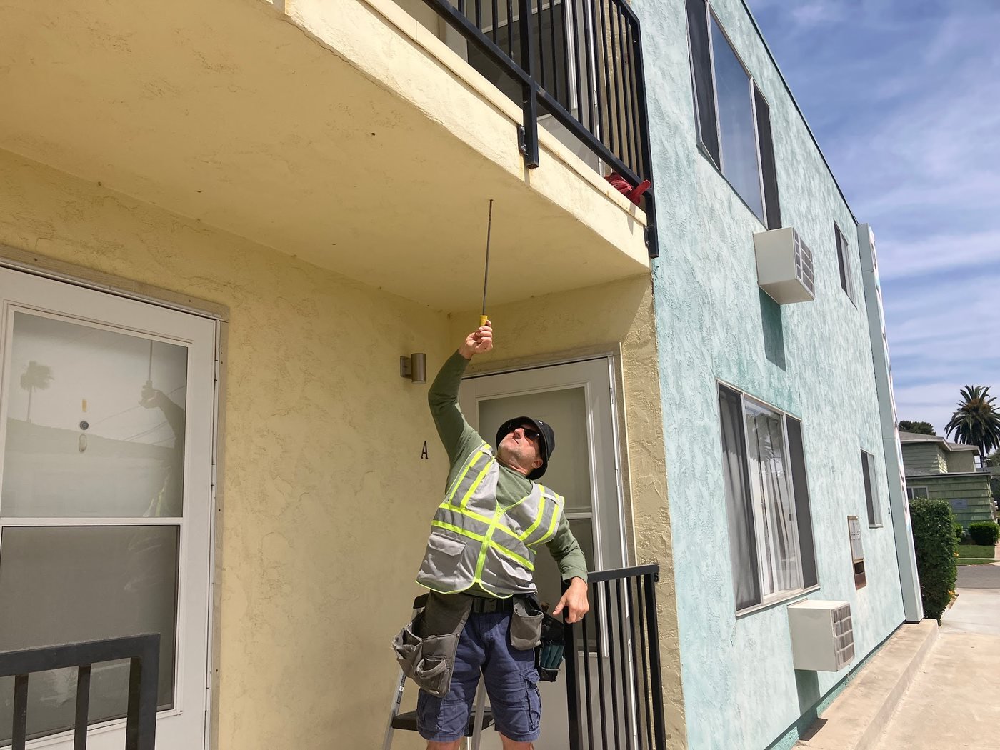 Inspector on a ladder using a probe tool to inspect the soffit edge beneath a second-floor balcony, with the full building facade visible